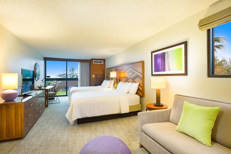 Spacious hotel room with two queen beds, ocean view balcony, modern furnishings, and sitting area with green accent pillow