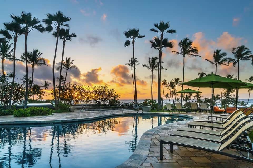 Illuminated pool at sunset with palm trees, green umbrellas, lounge chairs, and dramatic golden sky reflection