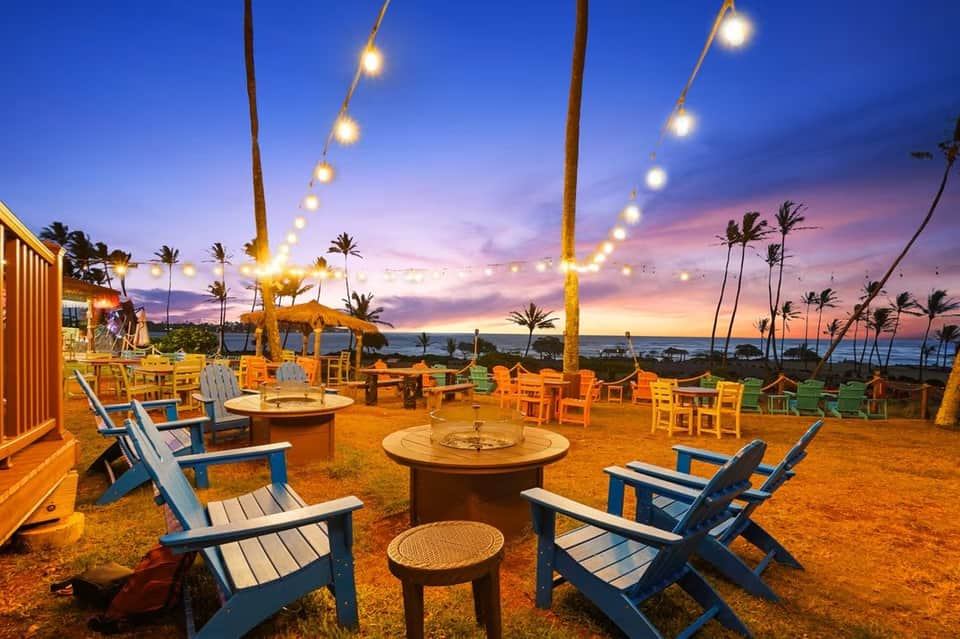 Beachfront fire pit lounge at dusk with Adirondack chairs, palm trees, and string lights overhead