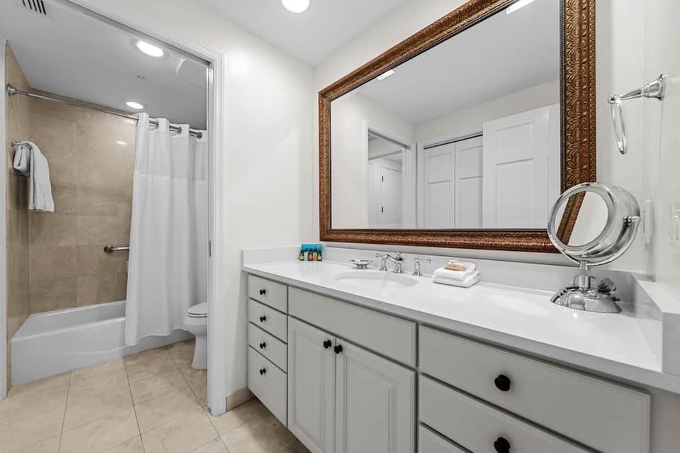 Clean white bathroom with double vanity, large mirror, shower/tub combination, and neutral tile flooring