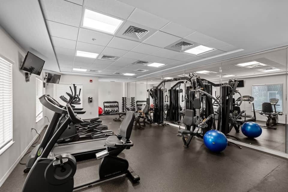 Bright fitness room with ellipticals, stationary bikes, weight equipment, blue exercise balls, and TV monitors