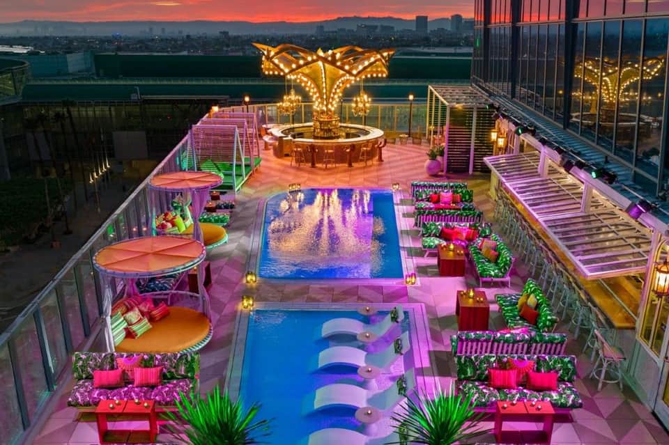 Rooftop pool complex with neon pink and purple lighting, illuminated structures, tropical landscaping at sunset