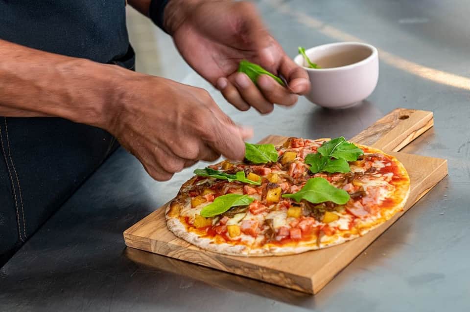 Person garnishing fresh pizza with basil on wooden board, with white cup and beverage nearby on table