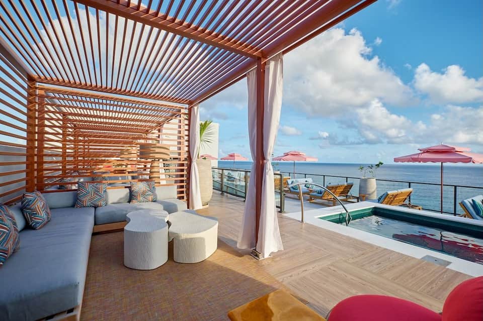 Modern covered deck with wooden pergola, gray sectional seating, white poufs, and pool with ocean horizon