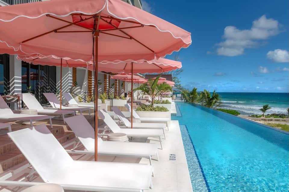Infinity pool overlooking beach with white lounge chairs under pink umbrellas, ocean and palm trees visible