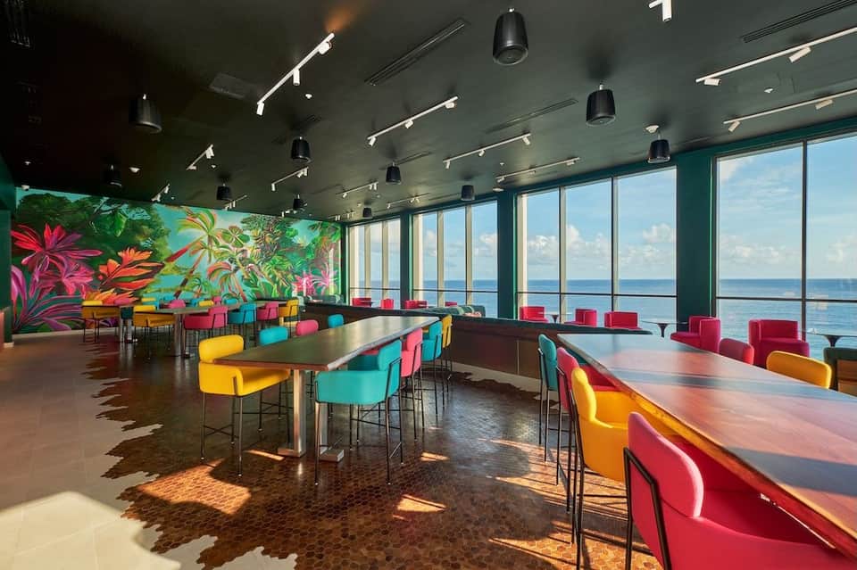 Modern waterfront lounge with colorful chairs, floor-to-ceiling ocean views, floral mural, and contemporary lighting fixtures