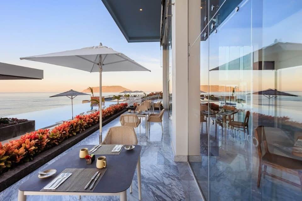 Waterfront dining area with glass walls, tables, umbrellas, and sunset views over calm water