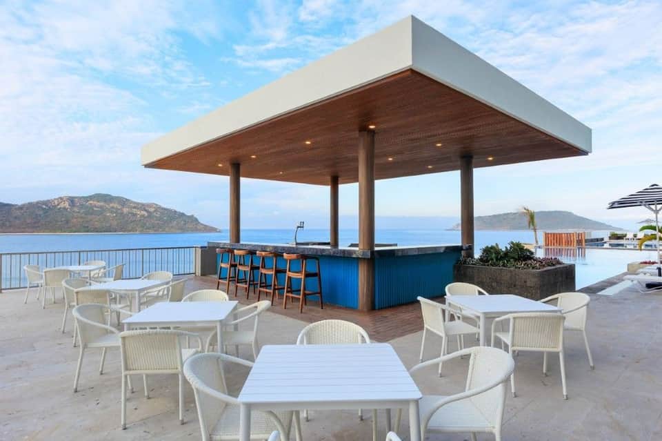 Beachfront bar pavilion with wooden ceiling, blue counter, and white dining tables overlooking Mediterranean sea and islands