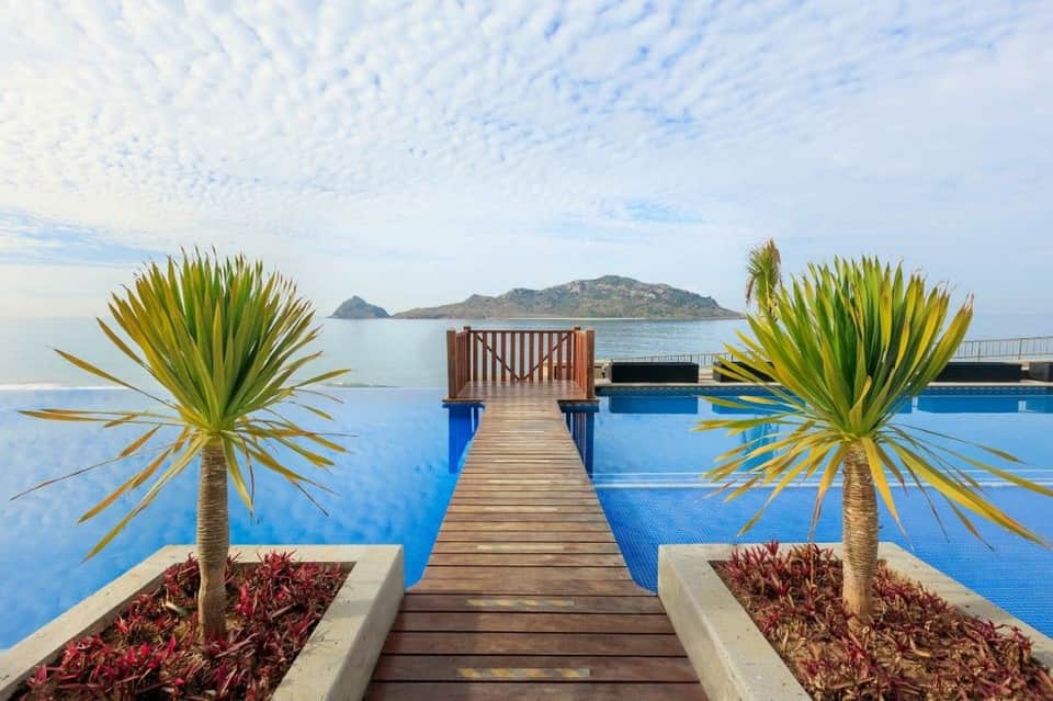 Wooden boardwalk with palm planters leading to infinity pool overlooking calm blue sea and rocky island landscape