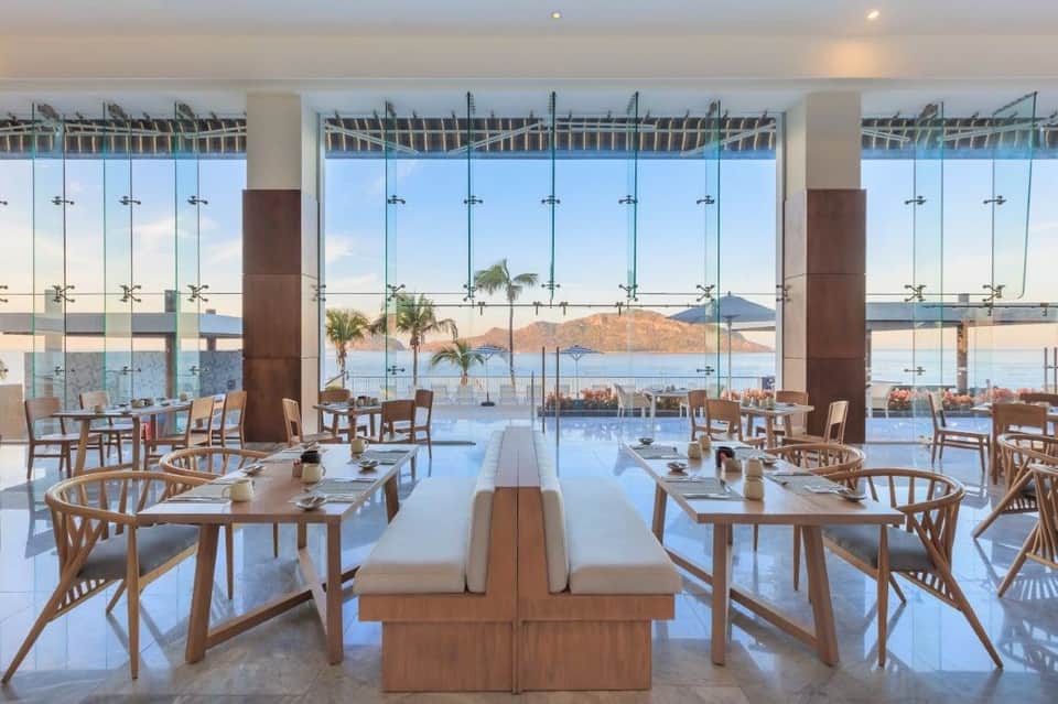 Elegant dining room with wooden chairs, round tables, floor-to-ceiling windows framing coastal mountain views at dusk