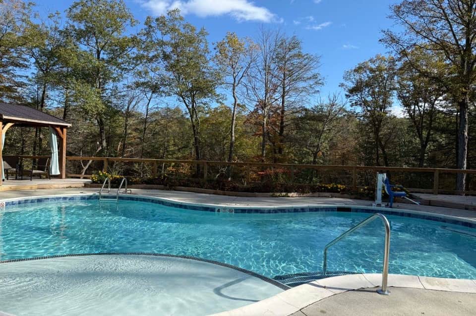 Crystal-clear resort pool with hot tub, wooden deck, gazebo, and autumn forest landscape.