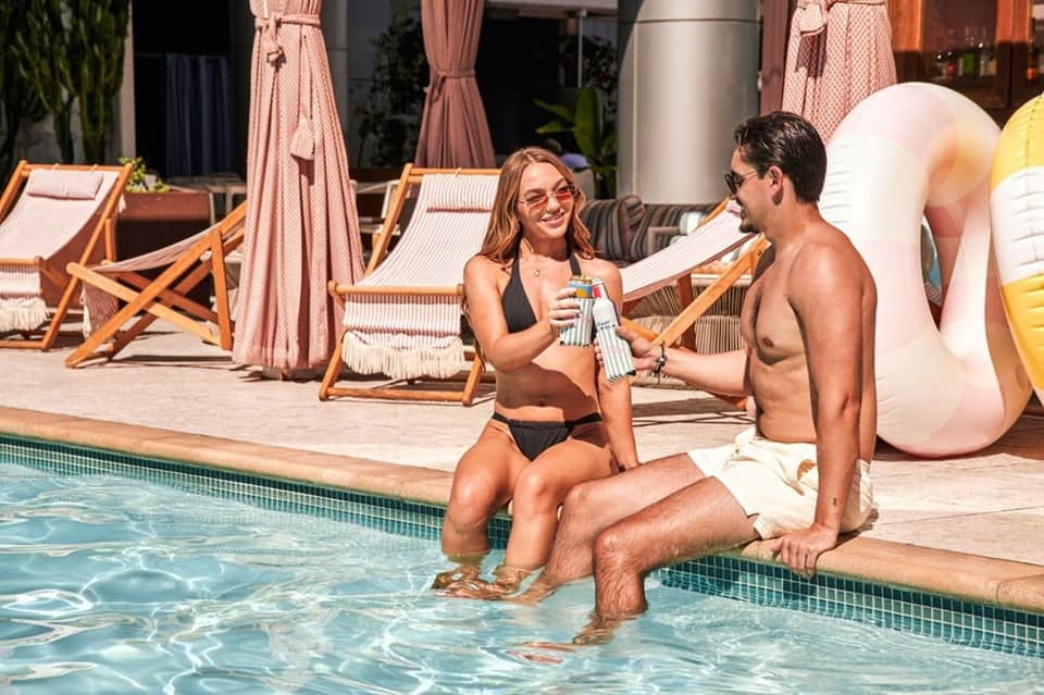 Two guests enjoying drinks by the resort pool with loungers and pink umbrellas in the background