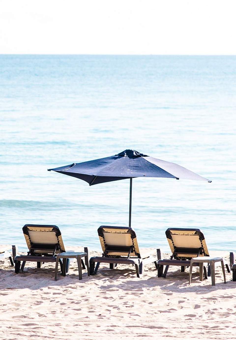Three lounge chairs with single blue umbrella on sandy beach facing calm turquoise water