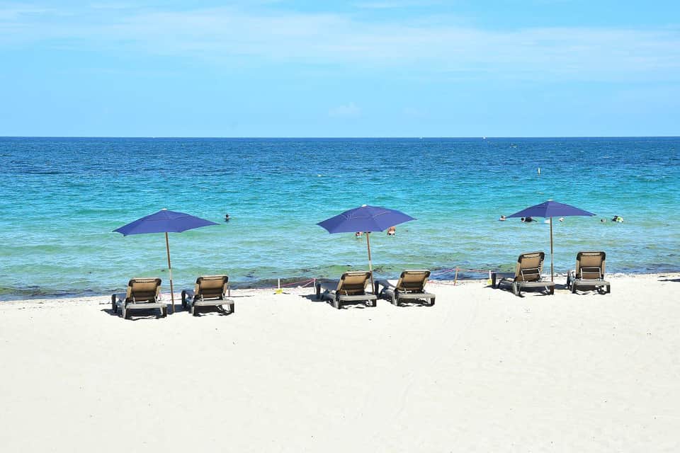 Multiple lounge chairs grouped under purple umbrellas on white sand beach with clear blue ocean