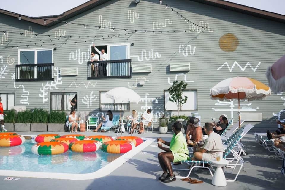 Modern resort pool area with colorful floats, guests lounging in chairs, and building with contemporary mural art