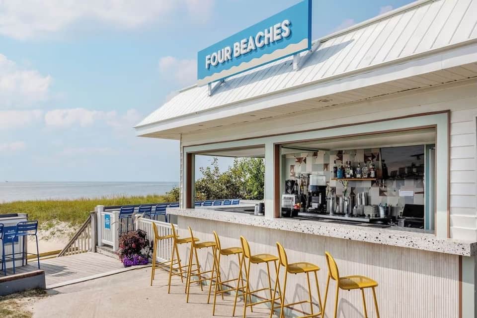 Four Beaches waterfront bar with service window, yellow bar stools, and beach access beyond