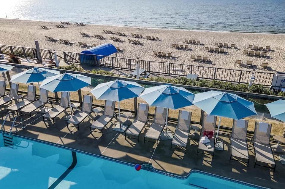 Beachfront pool area with blue umbrellas, lounge chairs, and sandy beach with sunbathers