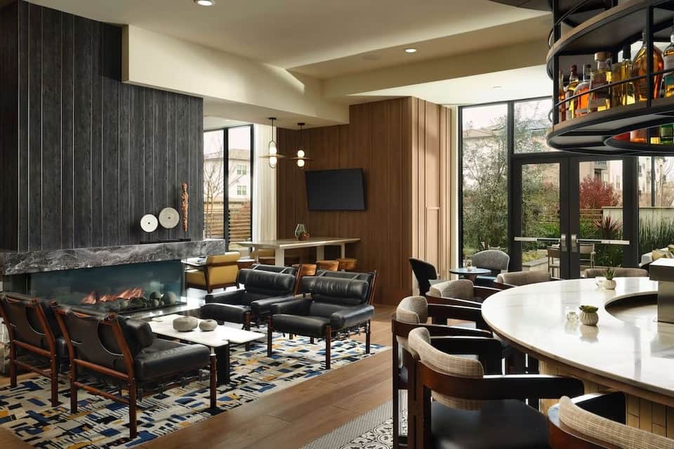 Luxury living room with fireplace, modern bar, leather seating, wood accents, and large windows overlooking landscape