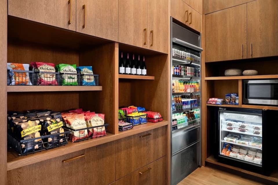 Hotel pantry with wooden cabinets, snack storage, beverage cooler, wine display, and refrigerated dessert case