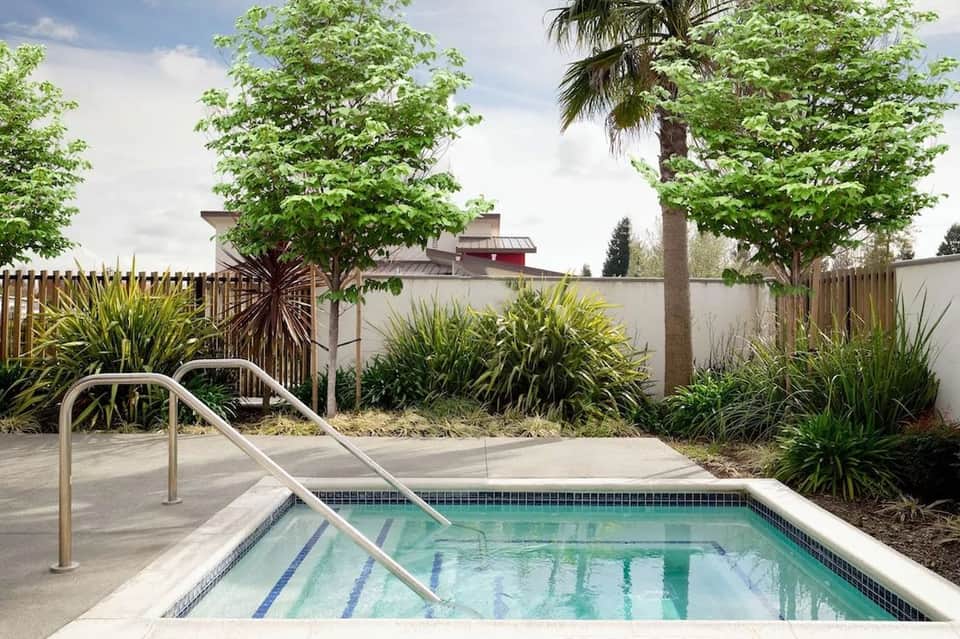 Small resort swimming pool with metal handrails, tile edging, white concrete deck, and tropical landscaping with palm trees