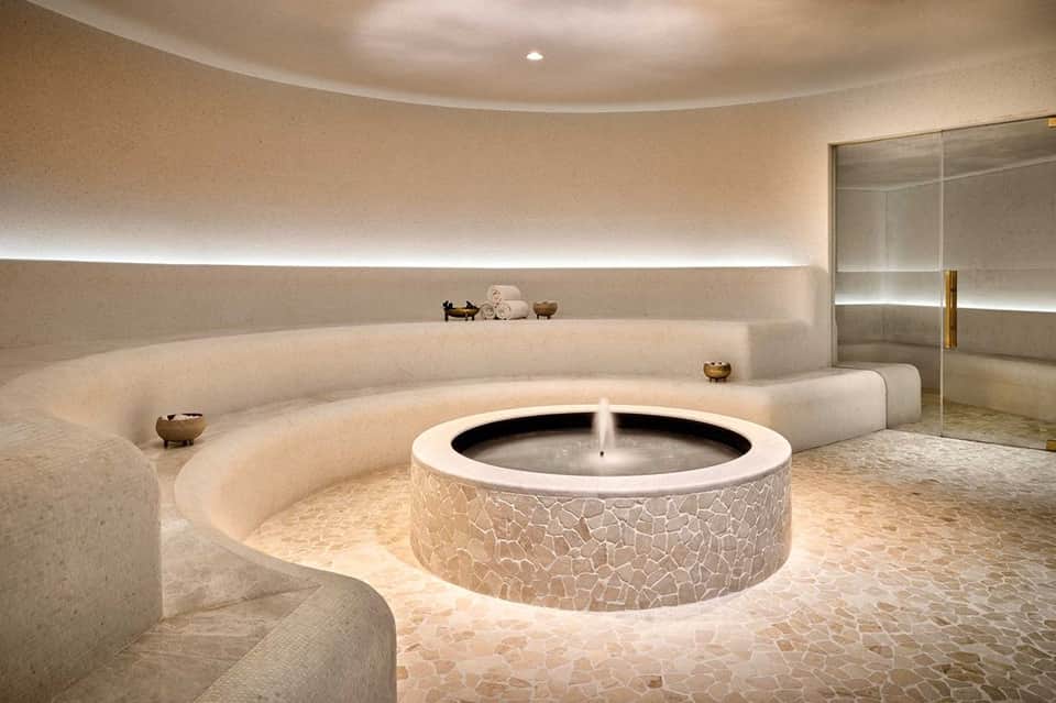 Luxurious circular spa with central fountain, curved seating, warm lighting, and glass shower enclosure