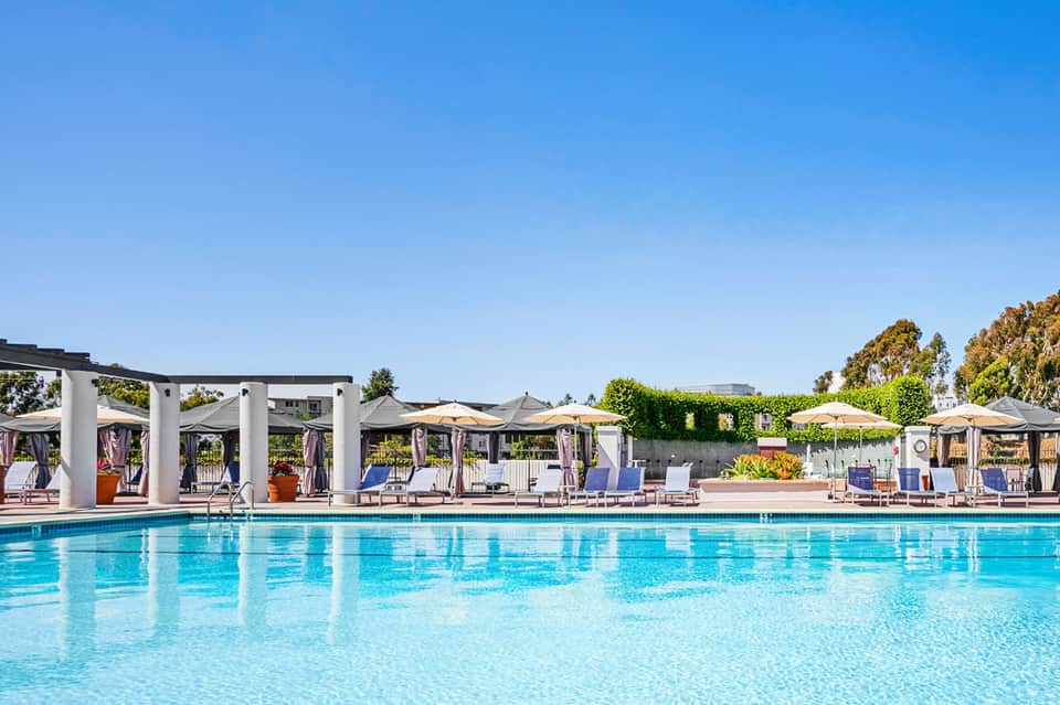 Resort outdoor pool with loungers, umbrellas, and white pergolas under clear blue sky