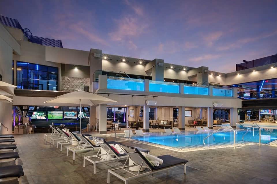 Upscale resort pool at dusk with lounge chairs, modern architecture, and lit buildings