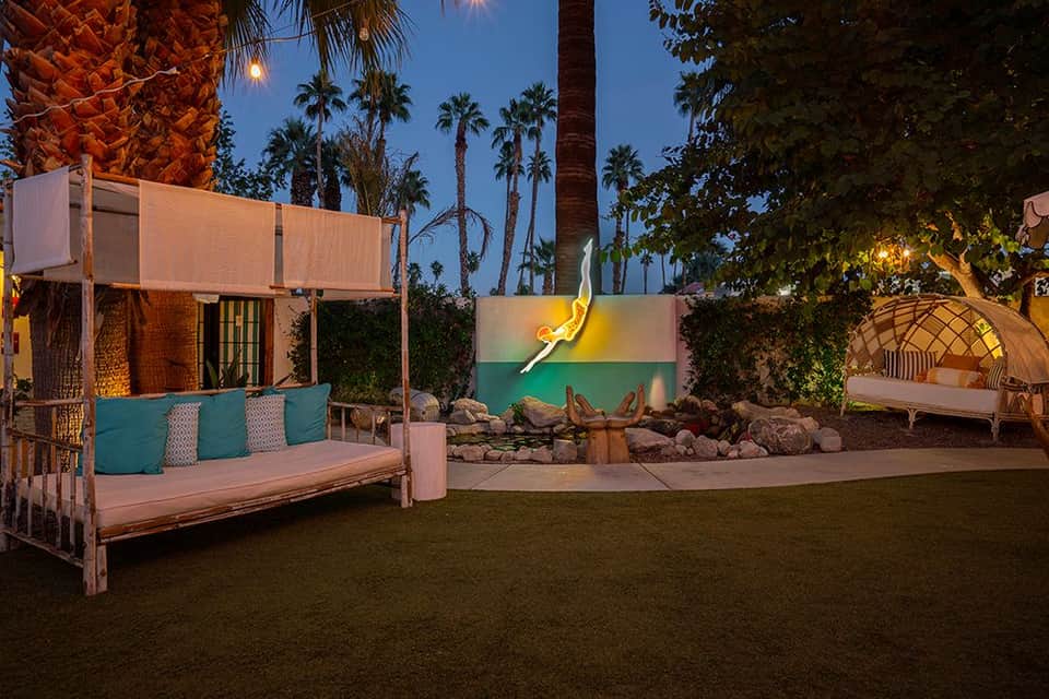 Nighttime resort courtyard with cushioned lounges, colorful wall art, and palm trees under blue sky