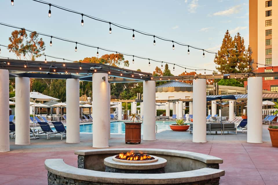 Resort pool area with fire pit, string lights, white columns, and lounge chairs at dusk