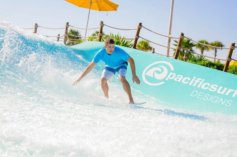Surfer riding artificial wave on shallow water ride with palm trees and yellow umbrella