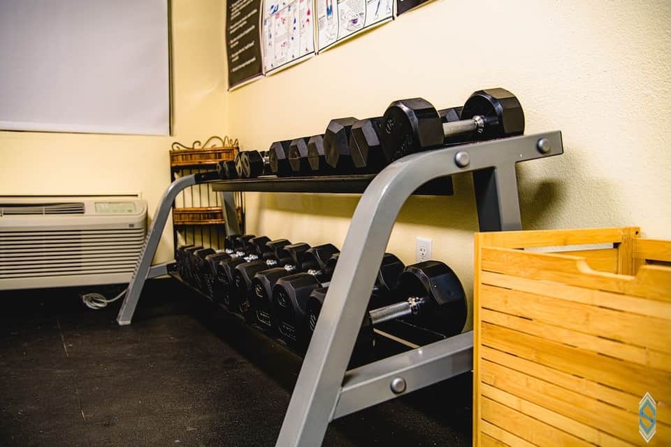 Exercise room displaying rack of dumbbells in various weights on gray metal stand with weight bench visible