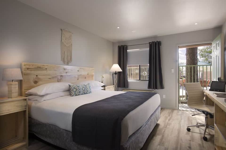 Modern hotel bedroom with queen bed, wooden headboard, desk, and glass door opening to balcony