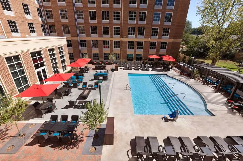 Outdoor pool area with red umbrellas, lounge chairs, and multi-story brick hotel building