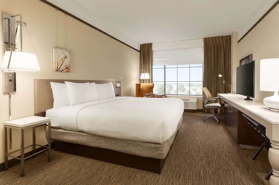 Modern hotel bedroom with queen bed, work desk, TV, and large windows with city view