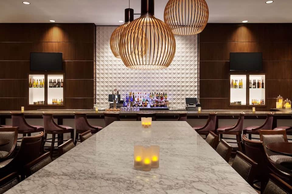 Modern hotel bar with marble countertop, wooden pendant lights, and illuminated bottle display