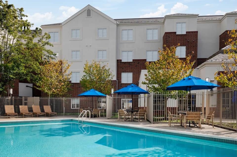 Outdoor swimming pool with blue umbrellas, lounge chairs, and multi-story hotel building with brick and beige facade