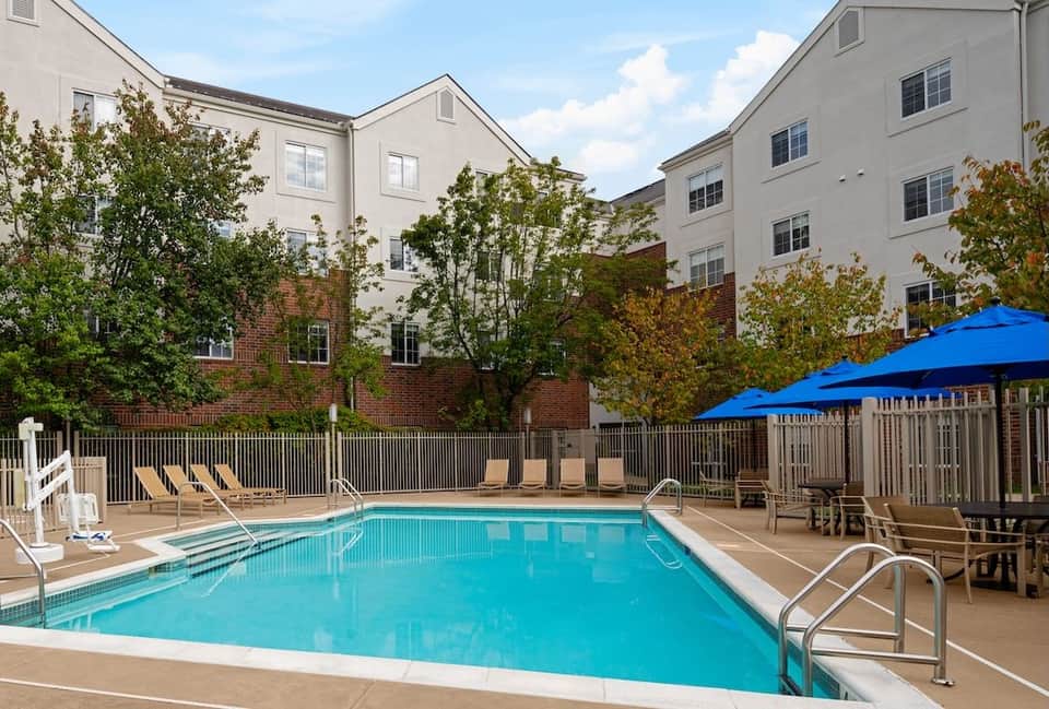 Resort swimming pool surrounded by lounge chairs, blue umbrellas, metal railings, and multi-story hotel buildings