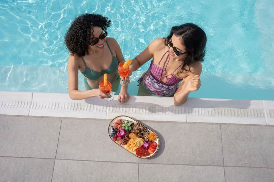 Two women in swimwear enjoying drinks by a resort pool with charcuterie board on deck