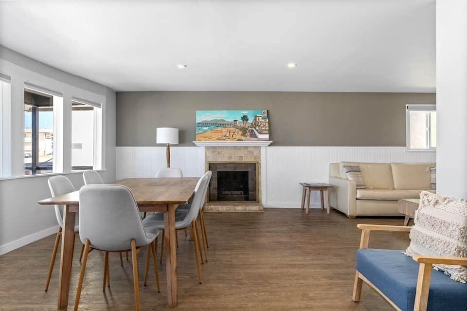 Spacious open-concept living area with dining table, fireplace, and coastal artwork, featuring white wainscoting and neutral tones