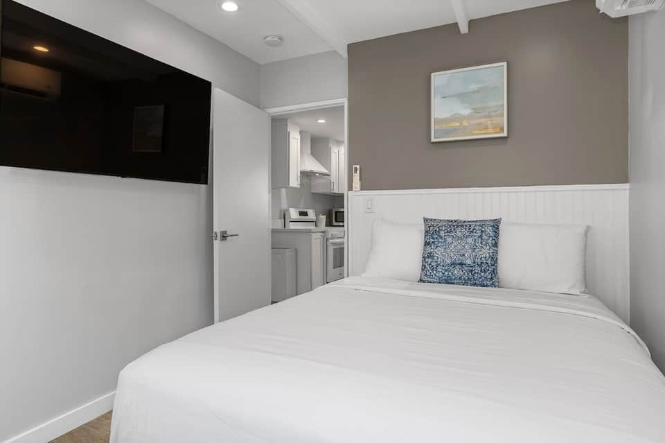 Compact bedroom with white bedding, beige and white wainscoting, and adjacent kitchenette