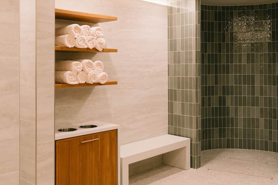 Spa bathroom with rolled white towels on wooden shelves, green tile shower area, and wooden vanity with white countertop