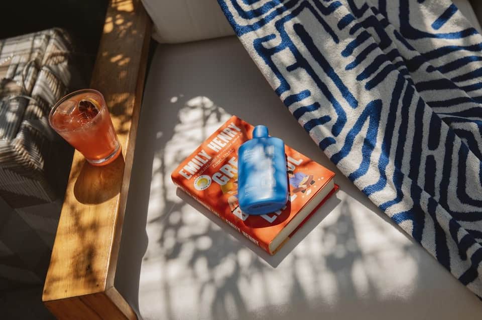 Sunlit table with coral cocktail, blue sunscreen bottle, orange magazine, patterned towel, and wooden furniture