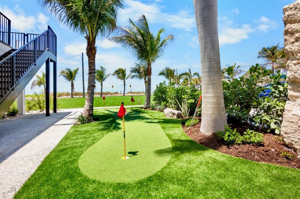 Putting green with red flag surrounded by palm trees and green grass near waterfront building
