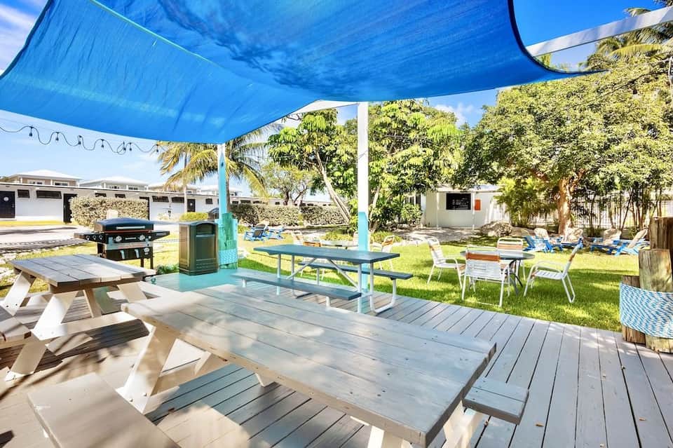 Outdoor picnic area with tables under blue shade sail, grill, and landscaped lawn