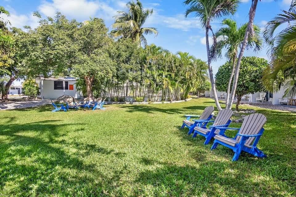 Multiple blue Adirondack chairs arranged on manicured lawn with palm trees and buildings