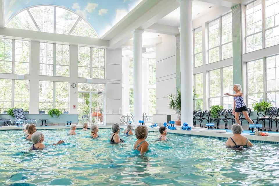 Indoor swimming pool with seniors exercising, large windows overlooking trees, instructor on deck