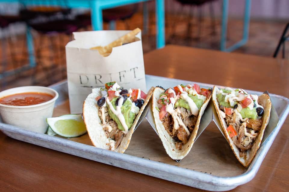Three filled tacos with shredded meat, lettuce, tomato, and toppings on wooden platter with lime