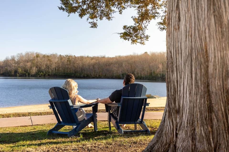 Couple in Adirondack chairs overlooking calm river with bare trees reflected in water