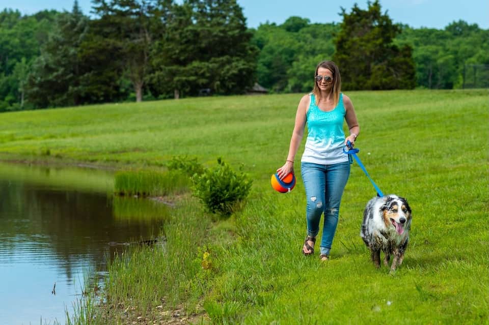Woman walking with dog along grassy field near pond with trees in background