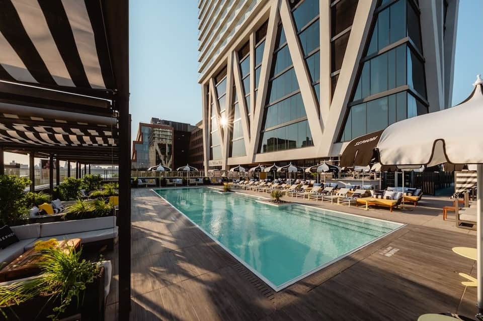 Modern rooftop pool with turquoise water, striped umbrellas, loungers, and contemporary city skyline backdrop